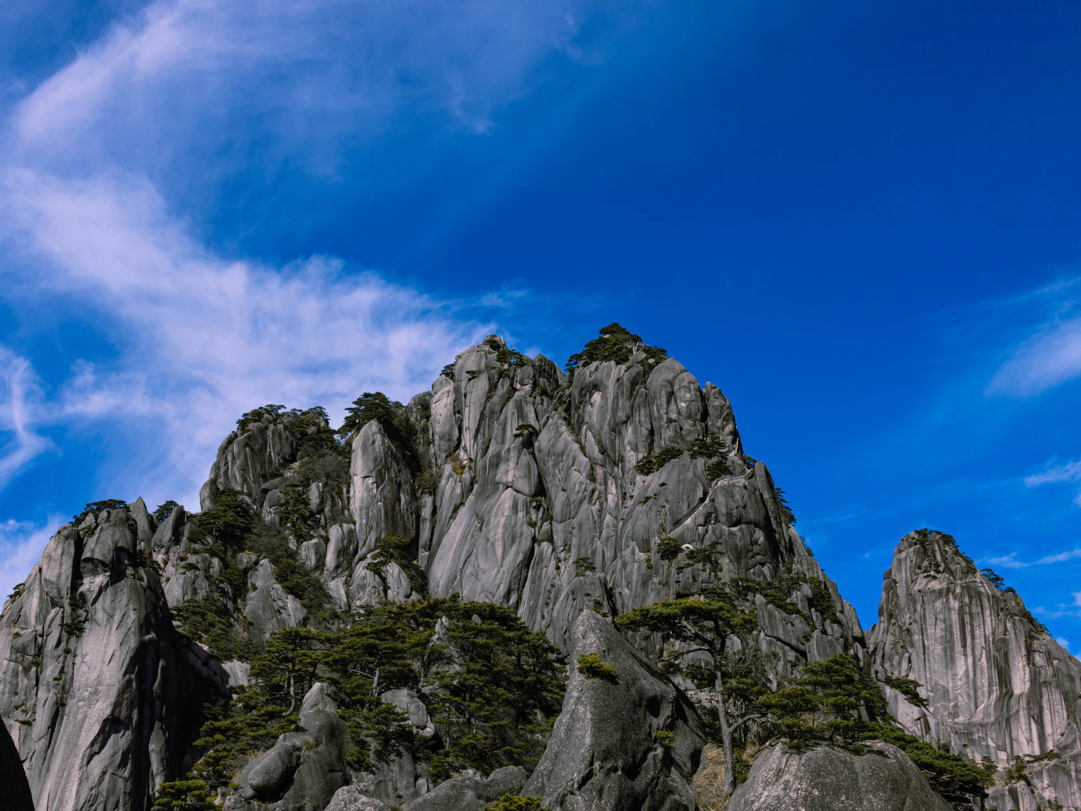 蓝天白云下的灰白色山峰，岩壁间生长着松树。